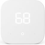 Amazon Smart Thermostat featuring a minimalist white rectangular design with a digital display, mounted on a modern home wall. The thermostat is showing the current temperature and energy-saving mode, emphasizing its integration with Alexa for easy voice control and smart home automation.
