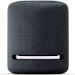 Amazon Echo Studio smart speaker with a sleek black cylindrical design, featuring a 360-degree audio setup and glowing blue LED ring, placed in a modern living room setting. The device is designed for hands-free control using Alexa, offering premium sound and seamless smart home integration.